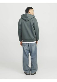Sudadera con capucha de tela verde oscuro y corte relajado, combinada con unos jeans de mezclilla azul claro y holgados, y zapatos negros, vista desde la parte trasera.