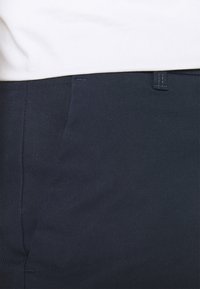 Pantalones azul marino oscuro con una textura suave, que presentan un bolsillo lateral y un detalle de costuras visibles. Combinados con una prenda superior de color claro.