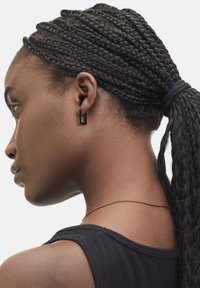 Schwarze geometrische Ohrringe aus Metall mit einem quadratischen Ausschnitt-Design. An einer Frau mit geflochtenem Haar, die ein schwarzes Oberteil trägt.