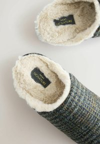 Grå-mønstrede slippers med en blød, beige imiteret pelsforing. Har et sort mærke med "NEXT SIGNATURE" i guldtekst indeni.