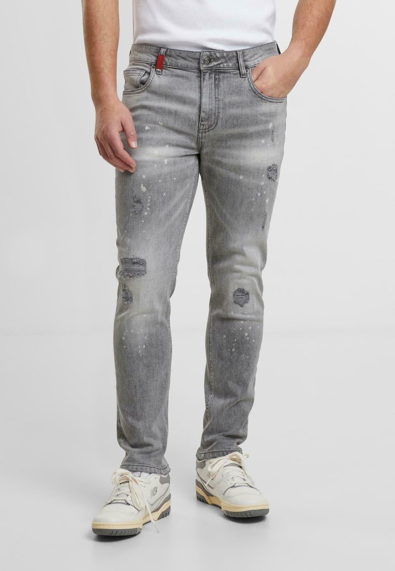 Jeans de mezclilla gris claro con un corte ajustado, con parches desgastados y detalles de salpicaduras de pintura. Combinados con zapatillas blancas.