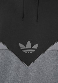 Svart och grå hoodie med Adidas trefoil-logotyp centrerad på bröstet och svarta dragband synliga ovanför logotypen.