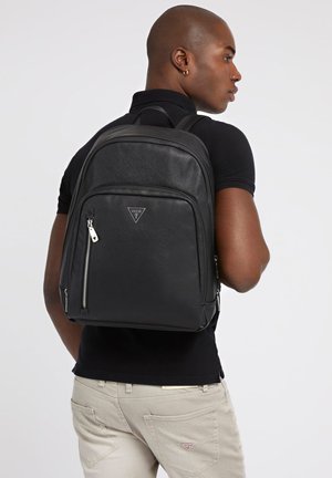Zaino in pelle nera con finitura texturizzata, tasca anteriore con zip e hardware in argento. Presenta un logo triangolare sul fronte.