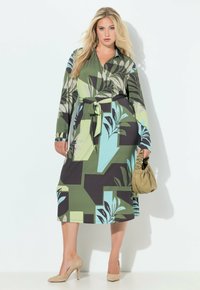 Wielokolorowa sukienka kopertowa z abstrakcyjnym wzorem liści w odcieniach zieleni, czerni i niebieskiego, długie rękawy i pas w tym samym wzorze; zestawiona z beżową torebką.