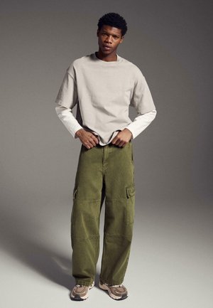 Tânăr în picioare, purtând cămăși cu mâneci lungi suprapuse în bej și alb, pantaloni cargo verde măsliniu și adidași bej pe un fundal neutru.