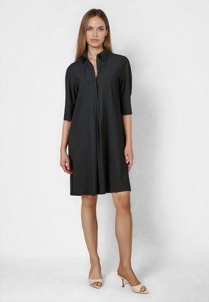 Woman standing wearing a knee-length black shirt dress with three-quarter sleeves and beige open-toe heeled mules against a plain background.