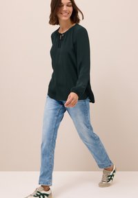 Blusa verde scuro a maniche lunghe con scollatura arricciata, abbinata a jeans a gamba dritta blu chiaro e sneakers grigie con dettagli verdi.