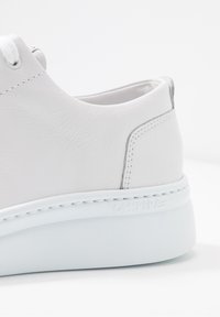 Zapatilla deportiva de cuero blanco con textura lisa, forma redondeada, suela blanca gruesa y costuras en contraste. Presenta un sutil logo de la marca en el lateral.