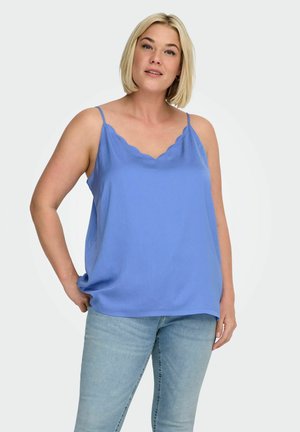 Kvinde med kort blondt hår iført en ærmeløs blå top med bølget kant og lyseblå jeans, stående foran en ensfarvet baggrund.