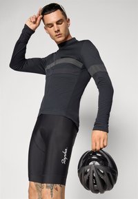 Black cycling jersey with long sleeves, featuring textured gray stripes and a zipper. Black cycling shorts with a logo. Black helmet held in hand.