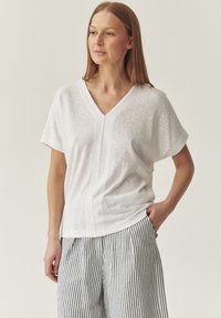 Top alb cu mânecă scurtă și decolteu în V, confecționat dintr-un material ușor, având o textură subtilă și un tiv drept, combinat cu pantaloni rayati.