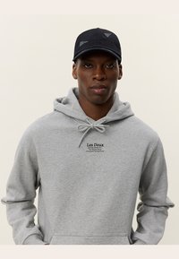 Grijze sweatshirt met trekkoorden en een kangoeroezak. Zwarte pet met geborduurde logo-accenten. Model kijkt recht vooruit met een neutrale uitdrukking.