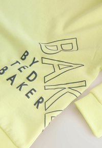 Jasnożółta bluza z charakterystycznym czarnym napisem "UPIEKŁO TED BAKER". Gładka tekstura z ribbami na mankietach.