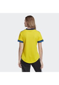 Gul sportstrøje med en tekstureret overflade, marinefarvet krave og blå detaljer på ærmerne, båret med sorte bukser. Har en buet kant.