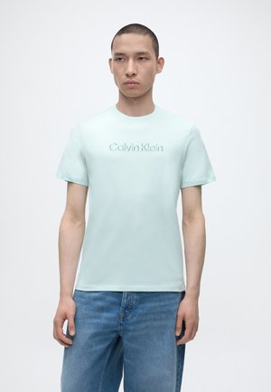 Νέος άνδρας με ανοιχτό μπλε μπλουζάκι Calvin Klein και μπλε τζιν, στέκεται μπροστά σε απλό λευκό φόντο.