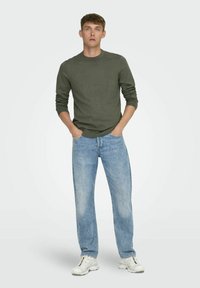 Pull en tricot vert avec col rond, associé à un jean droit bleu clair. Le mannequin se tient avec les mains dans les poches, portant des baskets blanches.