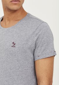 Tricou gri, cu mânecă scurtă, realizat dintr-un material moale, având guler rotund și un design brodat cu o palmier de culoare vișinie pe partea stângă a pieptului.