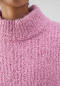 Rosa gebreide trui met een hoge, geribbelde kraag. De textuur is zacht en vachtachtig, met een subtiel patroon van variërende dikte in de stof.