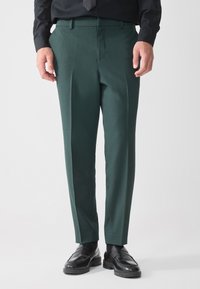 Grüne Anzughosen mit einem maßgeschneiderten Schnitt, die eine glatte Textur, Vorderfalten und einen Bund aufweisen. Kombiniert mit schwarzen Dressschuhen.
