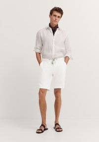 White linen shirt with rolled sleeves, paired with white shorts featuring an adjustable drawstring. Brown sandals complete the look.