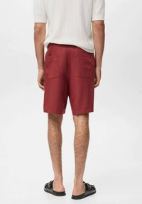Shorts rouges à coupe décontractée, dotés de deux poches arrière, fabriqués dans un tissu léger. Associés à des sandales noires et un haut en maille blanche texturée.