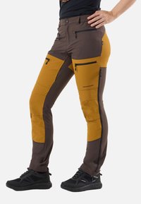 Brown and mustard slim-fit outdoor pants with multiple zip pockets worn with black sneakers and a black shirt.