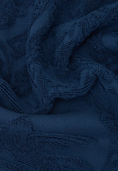 Tissu texturé bleu foncé avec des motifs géométriques en relief, montrant des plis et des fibres bouclées et douces.