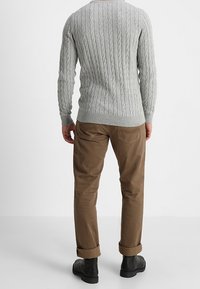 Šedý svetr s pletením, dlouhými rukávy, v kombinaci s béžovými chinos nohavicemi s ohrnutými lemy a černými kotníkovými botami, pohled zezadu.