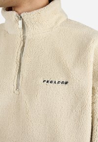 Kremowy polarowy sweter z wysokim kołnierzem i srebrnym zamkiem. Posiada czarne haftowane logo "PEGADOR" na klatce piersiowej. Miękka tekstura.
