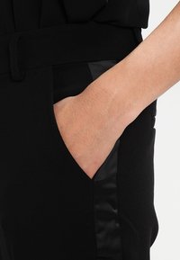 Svarta byxor i mjukt material med en satinpanel på sidan. Hand i fickan, vilket visar en ren design med en diskret midja.