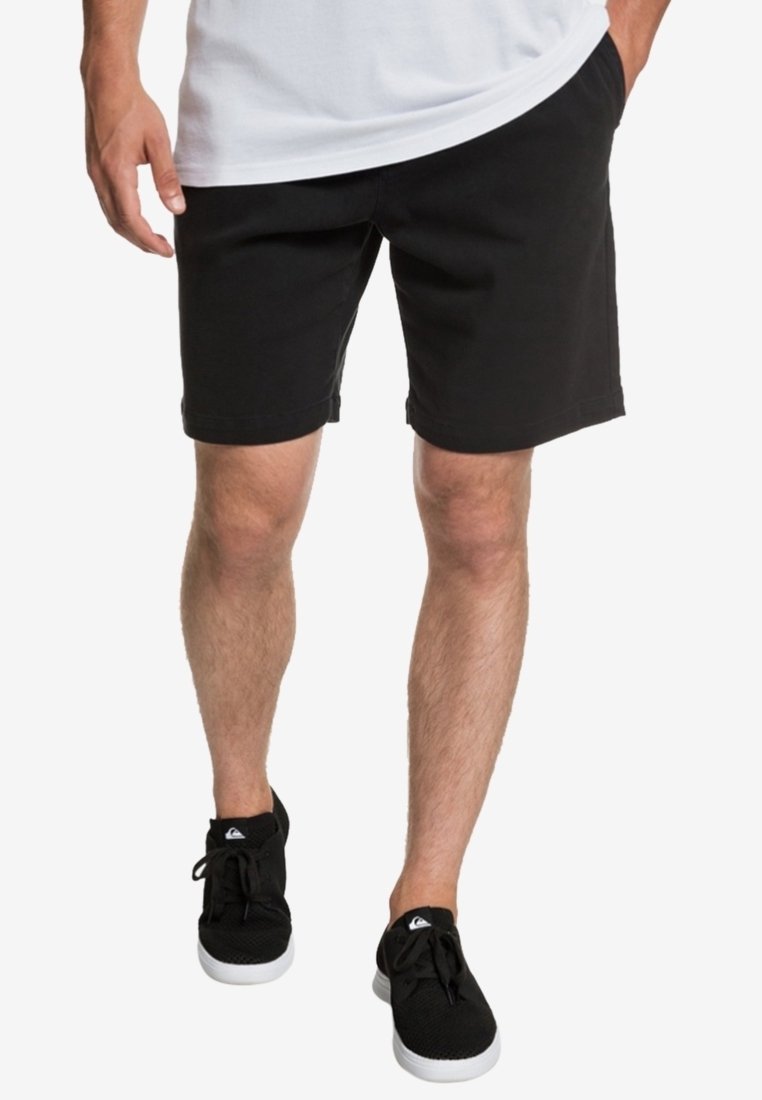 Homme portant un short noir, une chemise blanche et des baskets noires, marchant avec une main dans la poche sur fond blanc.