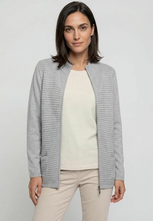 Vrouw met donker haar, gekleed in een lichtgrijze vest met rits over een beige shirt en beige broek, staand tegen een effen lichte achtergrond.
