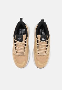 Beige sportcipők texturált hálós és bőr felsőrésszel, fekete belső béléssel, gumitalppal és rugalmas cipőfűzőkkel. Lacoste logó a nyelvén.