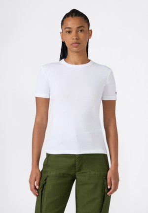 Mujer con cabello trenzado que lleva una camiseta blanca lisa de manga corta y pantalones cargo verde oliva, posando sobre un fondo blanco.