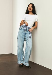 Weißes, kurzärmeliges Oberteil mit wellenförmigem Saum, kombiniert mit hellblauen Jeans mit weitem Bein und einem gestreiften Gürtel um die Taille. Schwarze Schuhe vervollständigen den Look.