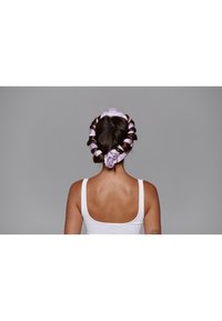 Des rouleaux de cheveux en satin violets sont stylisés en forme de cœur, maintenus par une chouchou lavande, sur un fond gris neutre.