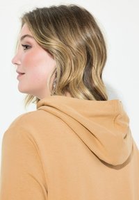 Haut à capuche marron clair avec une texture douce, présentant une vue de dos qui met en valeur la capuche et la encolure. Cheveux longs partiellement visibles.