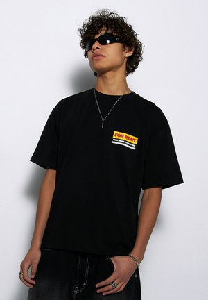 Czarny bawełniany t-shirt z krótkim rękawem, z kolorową grafiką "NA WYNAJEM" po lewej stronie klatki piersiowej. Noszony z szarymi dżinsami i akcesoriami.