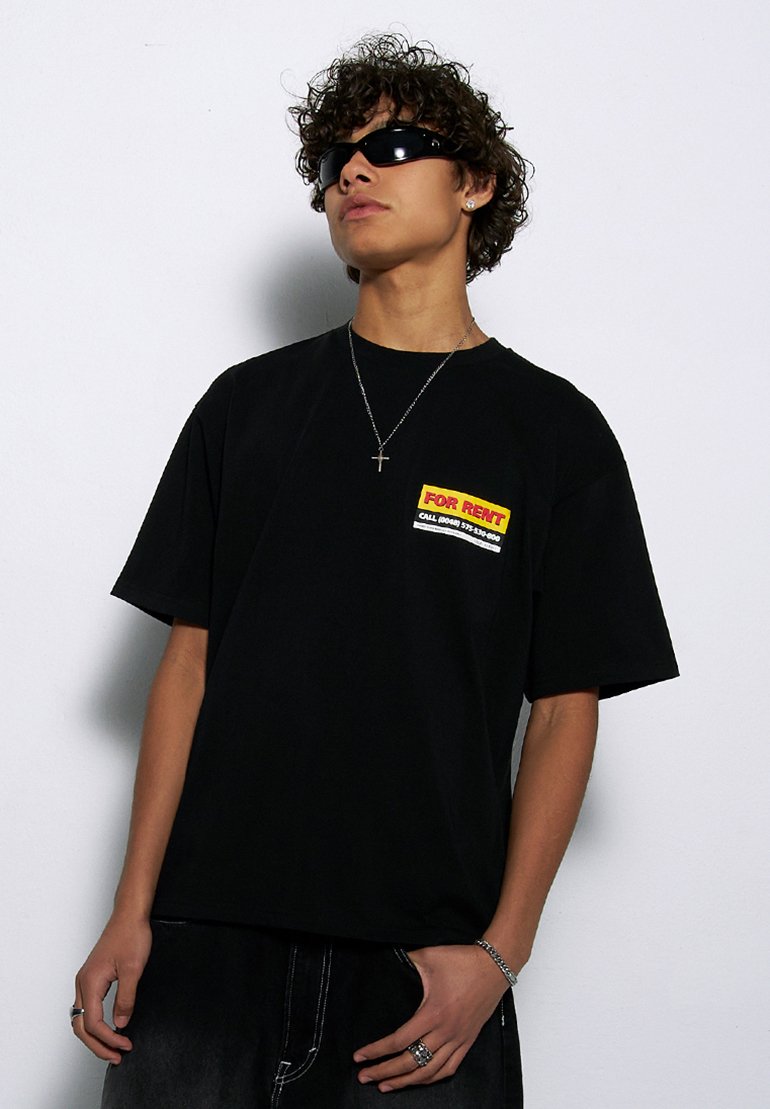 Czarny bawełniany t-shirt z krótkim rękawem, z kolorową grafiką "NA WYNAJEM" po lewej stronie klatki piersiowej. Noszony z szarymi dżinsami i akcesoriami.