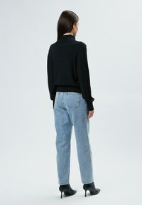 Schwarzer Strickpullover mit hohem Kragen, kombiniert mit hellblauen, hochgeschnittenen Jeans. Schwarze Stiefeletten runden das Outfit ab.