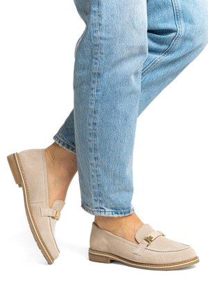 Beige Wildleder-Slipper mit goldfarbener Schnalle, getragen mit hochgekrempelten hellblauen Jeans bei überkreuzten Beinen.