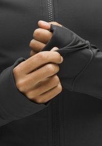 Manos ajustando los agujeros para el pulgar en una chaqueta negra con cremallera y mangas largas hecha de tela elástica.