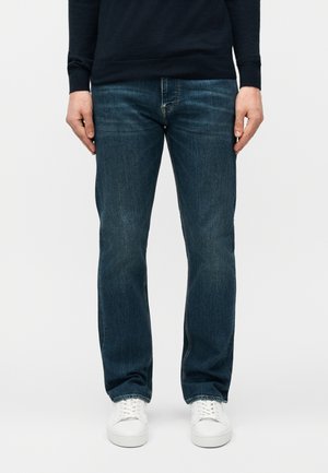 Man wearing dark blue straight-leg jeans and white sneakers, standing against a plain light background.