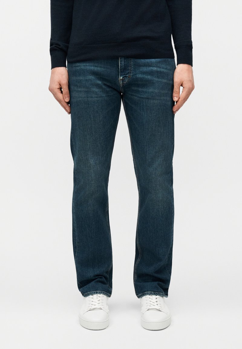 Άνδρας με σκούρο μπλε τζιν παντελόνι ίσιας γραμμής και λευκά αθλητικά παπούτσια, στέκεται μπροστά σε απλό ανοιχτόχρωμο φόντο.