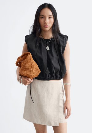 Jeune femme aux longs cheveux noirs porte un haut noir sans manches, une jupe beige, des bijoux en argent et tient un sac tressé marron.