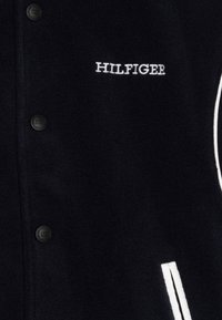 Granatowa polarowa kurtka z czarnymi guzikami, białymi akcentami i haftem "HILFIGER" w białym kolorze na przodzie. Prosty, czysty fason.