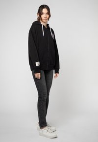 Zwarte zip-up hoodie met een capuchon, voorzien van een bloemenborduurontwerp. Gepaard met grijze skinny jeans en witte sneakers. Zachte stoffen textuur.