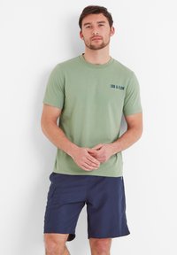 Green cotton t-shirt with "EBB & FLOW" printed in dark blue. Paired with navy shorts. Simple crew neck and short sleeves.