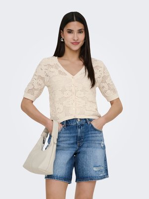 Kvinde med langt mørkt hår iført en beige strikket kortærmet knapbluse, denimshorts, sølv øreringe, der holder en lysebeige håndtaske.
