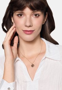 Złoty naszyjnik z okrągłym wisiorkiem z motywem drzewa. Modelka ubrana w białą, teksturowaną bluzkę, dotyka szyi.